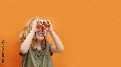 A cute little curly-haired girl looks through binoculars on monochrome background in studio. Copy space. The concept of travel and recreation, vacations and adventures, entertainment with child