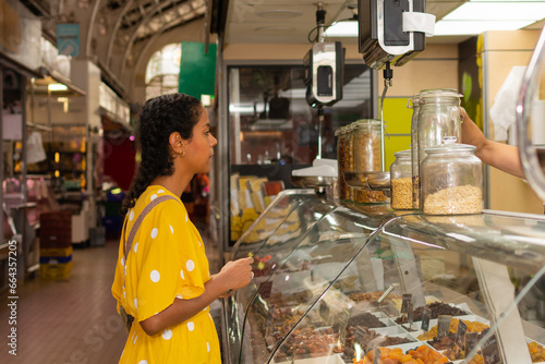 A young Hispanic woman in a yellow polka dot dress shopping for groceries in front of a display case filled with nuts and olives