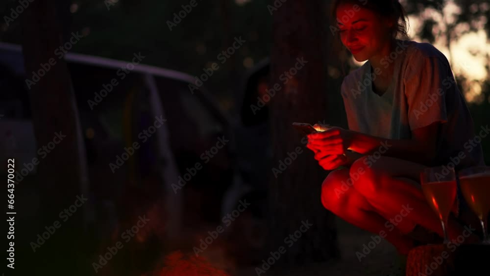 Hipster girl using smartphone sitting next to bonfire in forest camping at night. Woman hiking alone. Active leisure and lifestyle. People in woods. Tourist woman in forest campsite. Hipster girl using smartphone sitting next to bonfire in forest camping at night. Woman hiking alone. Active leisure and lifestyle. People in woods. Tourist woman in forest campsite.