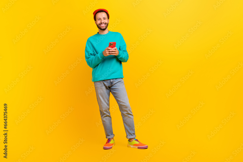 Fototapeta premium Full size photo of optimistic guy wear teal sweatshirt red hat hold smartphone in hands read email isolated on yellow color background