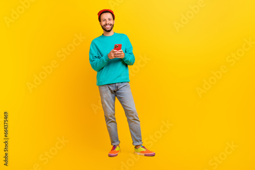 Full size photo of optimistic guy wear teal sweatshirt red hat hold smartphone in hands read email isolated on yellow color background