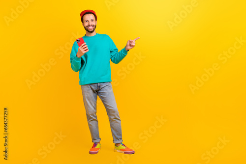 Full size photo of handsome guy wear teal sweatshirt hold smartphone indicating at offer empty space isolated on yellow color background © deagreez