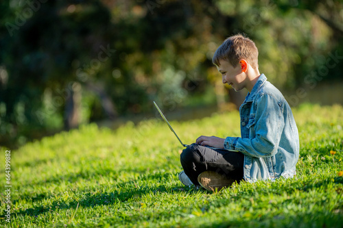 Teen Boy studying online with a computer  outdoors.  Smiling schoolboy with a laptop sits on a green lawn. Young cheerful school child sitting and typing on computer in the park. Looking to the screen