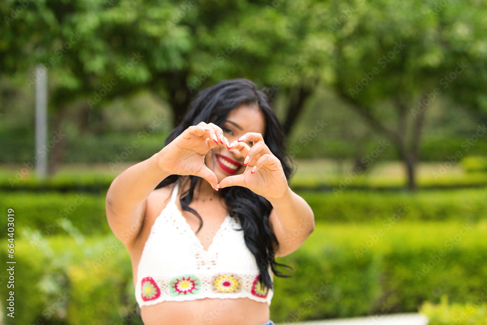 Obraz premium Young and beautiful brunette woman and latin, making a heart shape with her hand and fingers smiling looking through the sign. Concept friendship and love. 14 of february valentine's day.