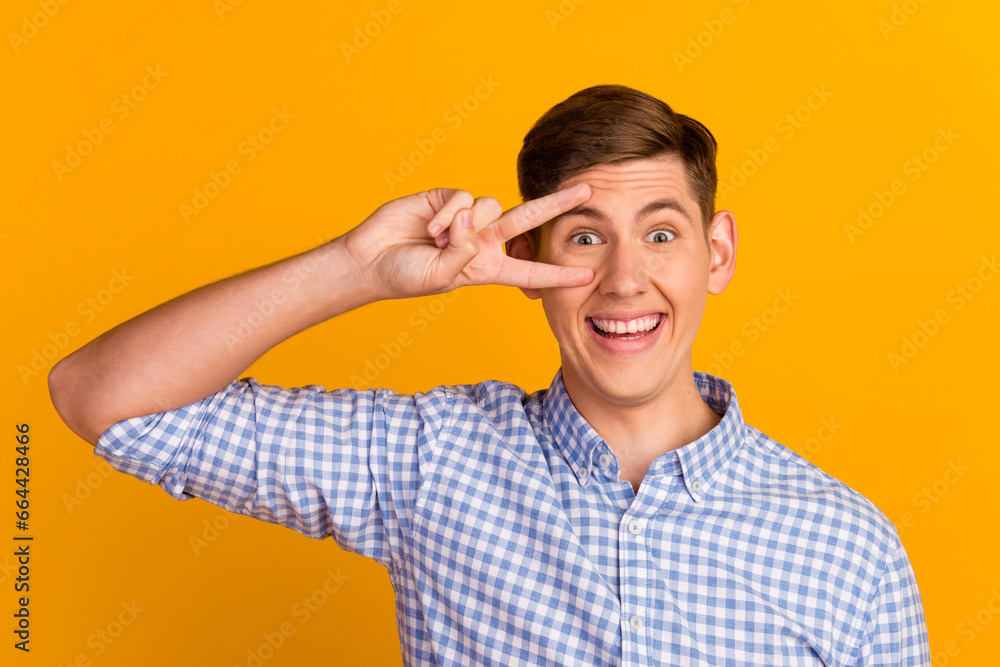 Photo of young smiling good mood guy showing v-sign near eye flirty mood isolated on yellow color background Photo of young smiling good mood guy showing v-sign near eye flirty mood isolated on yellow color background
