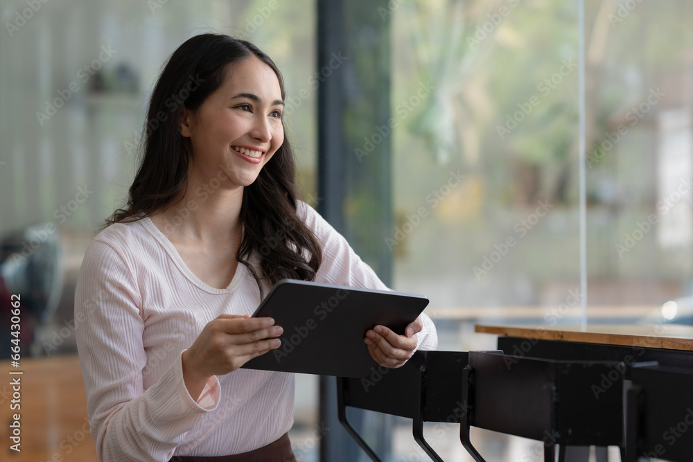 Portrait of a young happy Asian woman holding a digital tablet and looking away while standing in a cafe.