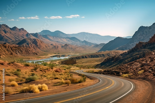 Curving asphalt road or highway leading through a picturesque desert landscape, framed by jagged mountains, with a winding river shimmering in the distance under a vast sky.