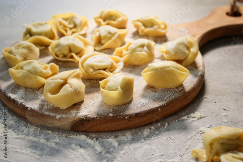 Raw traditional ravioli tortellini on wooden plate, grey table with flour