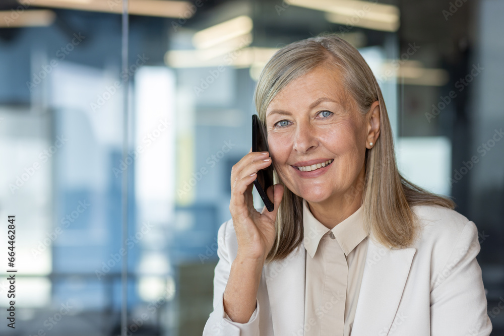 Close-up portrait of a smiling senior business woman in a suit sitting at a desk in the office and talking on a mobile phone. Close-up portrait of a smiling senior business woman in a suit sitting at a desk in the office and talking on a mobile phone.