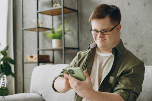 Young man with down syndrome wear glasses casual clothes use mobile cell phone chatting sits on grey sofa couch stay at home flat rest spend free time in living room Genetic disease world day concept photo