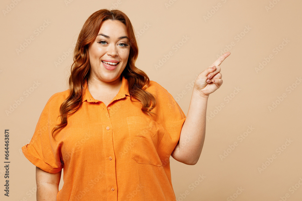 Young cheerful ginger chubby overweight woman wear orange shirt casual clothes point index finger aside indicate on area isolated on plain pastel beige background studio portrait. Lifestyle concept. Young cheerful ginger chubby overweight woman wear orange shirt casual clothes point index finger aside indicate on area isolated on plain pastel beige background studio portrait. Lifestyle concept.