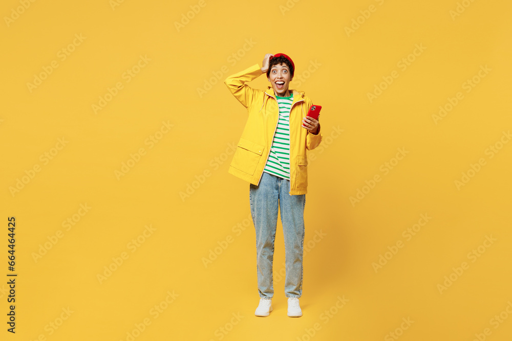 Full body young woman wearing waterproof raincoat outerwear red hat hold use mobile cell phone isolated on plain yellow background studio portrait. Outdoors lifestyle wet fall weather season concept. Full body young woman wearing waterproof raincoat outerwear red hat hold use mobile cell phone isolated on plain yellow background studio portrait. Outdoors lifestyle wet fall weather season concept.