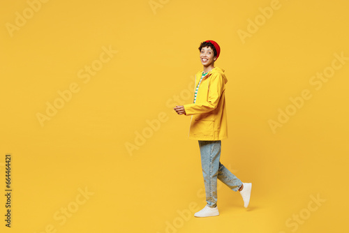 Full body side view young smiling happy woman wear waterproof raincoat outerwear red hat walk go isolated on plain yellow background studio portrait Outdoors lifestyle wet fall weather season concept