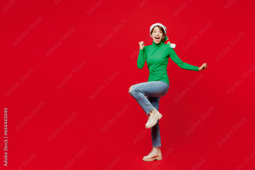 Full body young woman wear green turtleneck Santa hat posing do winner gesture celebrate clench fist say yes isolated on plain red background Happy New Year 2024 celebration Christmas holiday concept