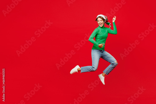 Full body side view young woman in green turtleneck Santa hat posing jump high pov play guitar hand gesture isolated on plain red background Happy New Year 2024 celebration Christmas holiday concept © ViDi Studio