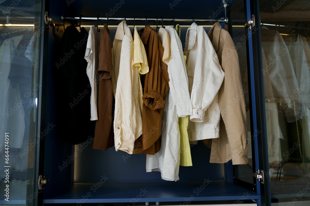 Horizontal image of clothes - dresses, shirts and t-shirts hanging in a blue backlit closet. The concept of tidy wardrobe storage, order and style
