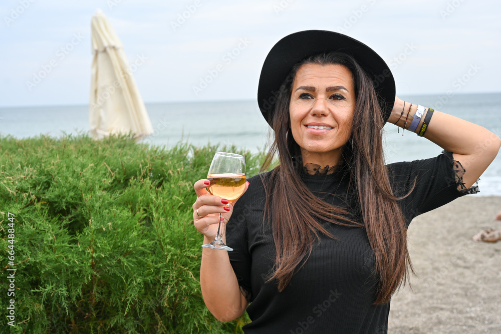 Fototapeta premium Portrait of Young Beautiful Smiling happy woman standing with glass of wine against sea during the winter in leather jacket and hat .