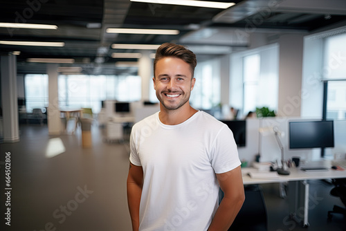 A smiling young, handsome man of European origin, with a stylish haircut, and white teeth, standing in a blank white t-shirt on a blurred background of a large empty office.