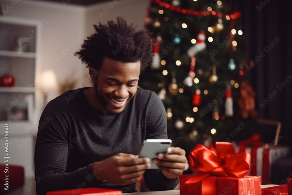 Young afro american male ordering a christmas present on phone sitting next to a christmas tree. Smiling happy young african male holding phone. Young afro american male ordering a christmas present on phone sitting next to a christmas tree. Smiling happy young african male holding phone.