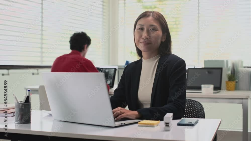 Medium portrait shot of female office worker texting messages to clients on laptop and looking at camera Medium portrait shot of female office worker texting messages to clients on laptop and looking at camera