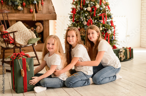 Three pretty young girls in the living room are sitting on the floor near the Christmas tree. Christmas, New Year concept. © Glebsterr