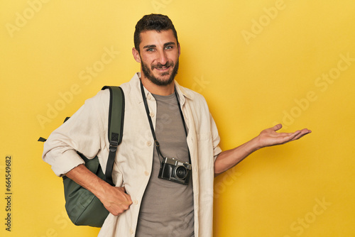 Young Hispanic travel photographer poses showing a copy space on a palm and holding another hand on waist.