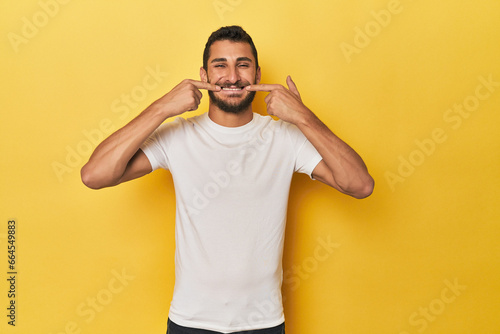 Young Hispanic man on yellow background smiles, pointing fingers at mouth.