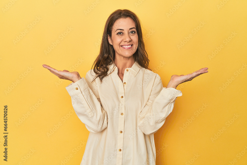 Middle-aged caucasian woman on yellow makes scale with arms, feels happy and confident. Middle-aged caucasian woman on yellow makes scale with arms, feels happy and confident.