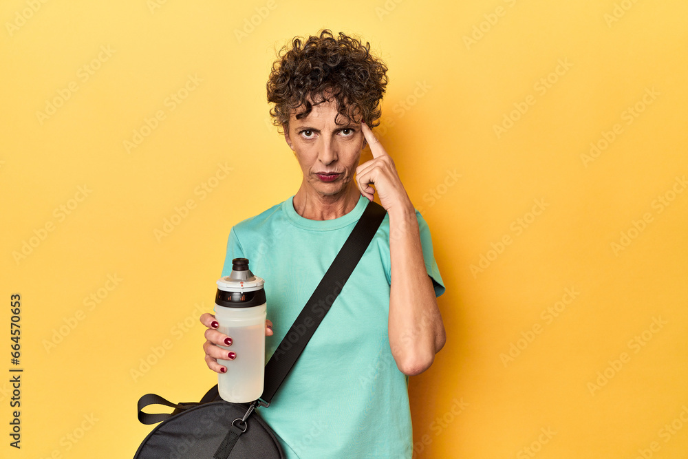Sportswoman with gym bag & water on yellow studio pointing temple with finger, thinking, focused on a task. Sportswoman with gym bag & water on yellow studio pointing temple with finger, thinking, focused on a task.