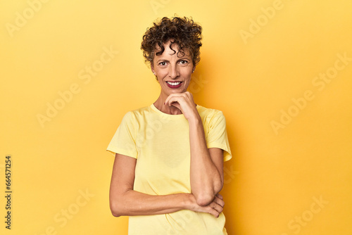 Mid-aged caucasian woman on vibrant yellow smiling happy and confident, touching chin with hand.