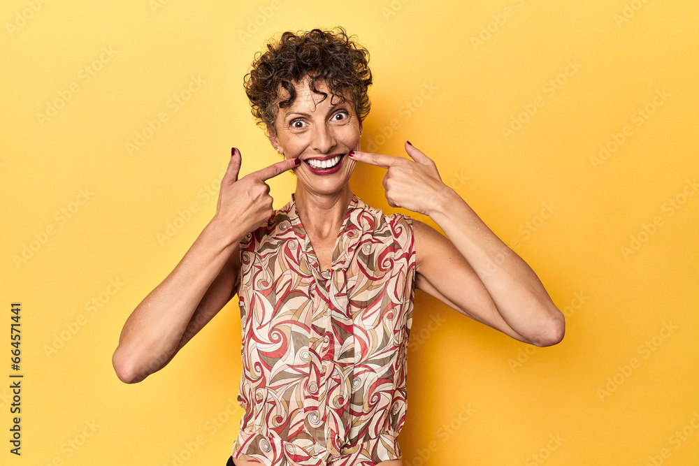 Mid-aged caucasian woman on vibrant yellow smiles, pointing fingers at mouth. Mid-aged caucasian woman on vibrant yellow smiles, pointing fingers at mouth.