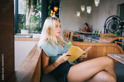 Contemplative Caucasian student with education textbook for organization planning pondering on test information while doing homework in cafe interior, pensive blonde woman learning and studying