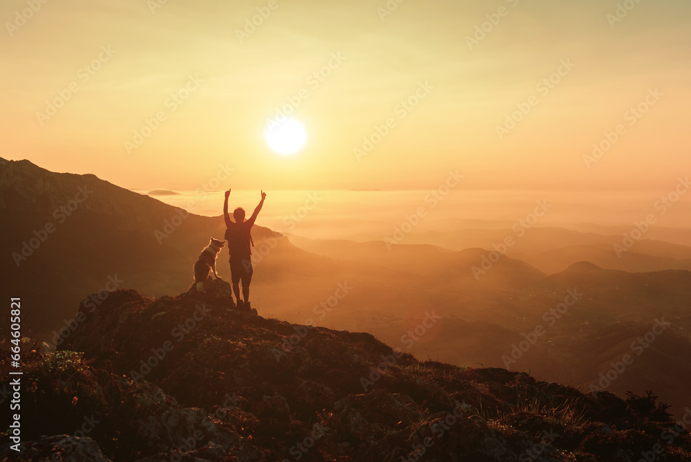 silhouette of a mountaineer man raising his arms and contemplating the sunset after hiking in the mountains with his border collie breed dog. outdoor sports and adventure