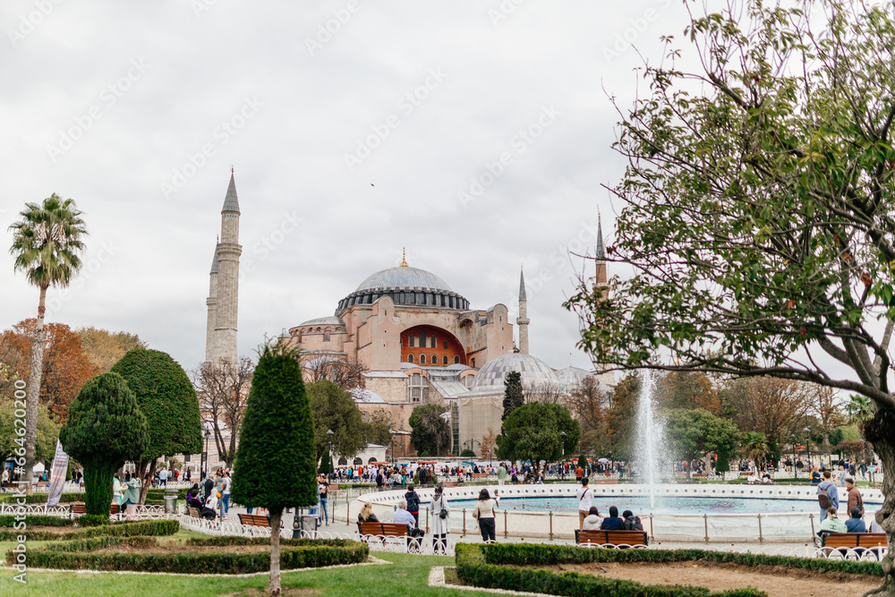 Ancient architecture in the middle of a square with a fountain, lots of tourists. City landscape with an old temple on an autumn cloudy day. Hagia Sophia, Fatih, Istanbul, Türkiye - October 16, 2023