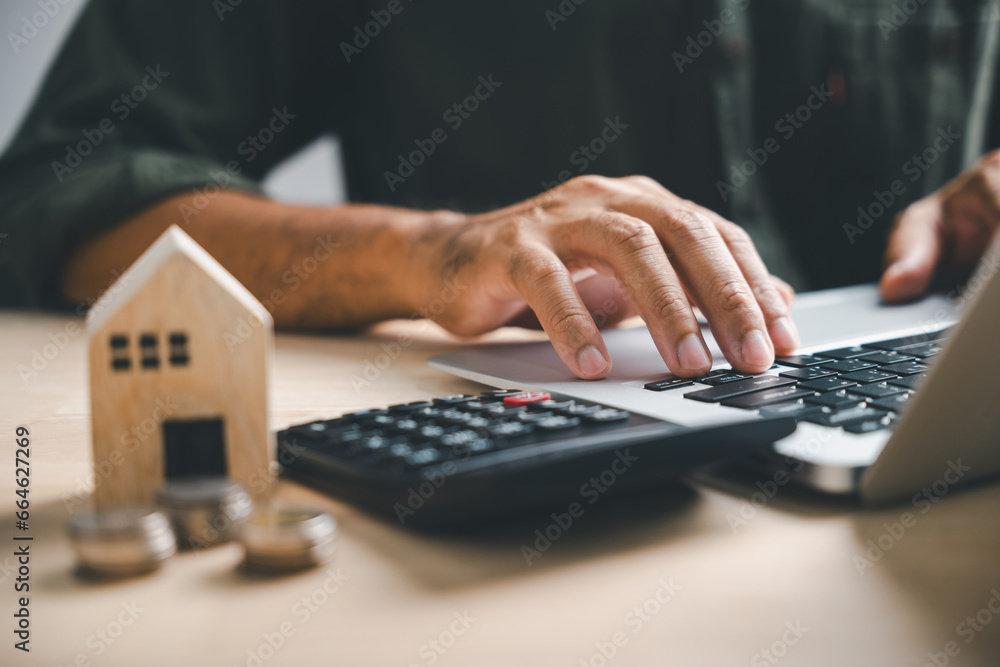 Hand presses calculators, pondering home refinance. Wooden house model, buy or rent note on desk. Saving for property purchase, mortgage payment strategy. Tax, credit analysis for financial success. - obrazy, fototapety, plakaty 