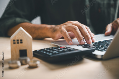 Hand presses calculators, pondering home refinance. Wooden house model, buy or rent note on desk. Saving for property purchase, mortgage payment strategy. Tax, credit analysis for financial success. photo