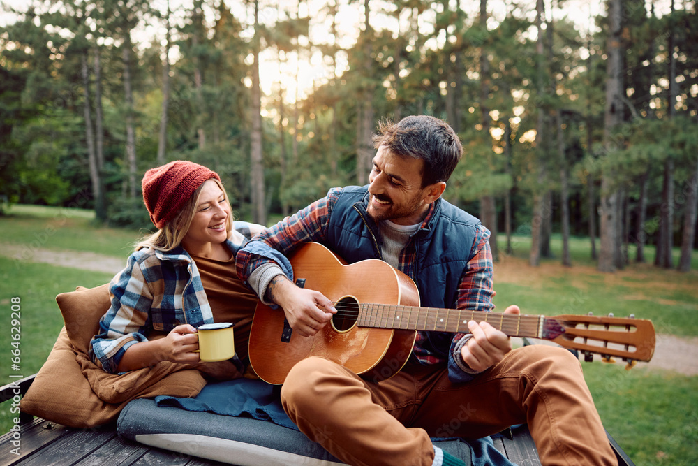 Happy man plays acoustic guitar to his girlfriend while camping in woods. Happy man plays acoustic guitar to his girlfriend while camping in woods.