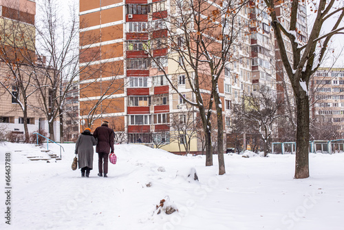 An old man and an old woman are walking along a snowy road to a high-rise building in a residential area of the city in winter © Maryna