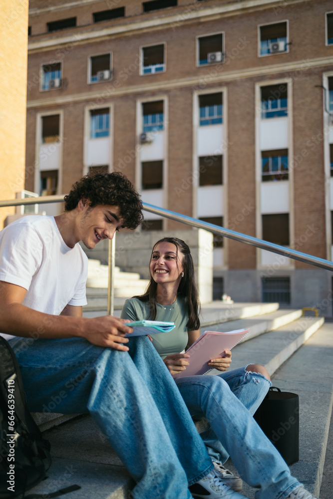 Young and happy college students with casual clothes sitting on stairs outdoors at university and talking while talking and studying with notes Young and happy college students with casual clothes sitting on stairs outdoors at university and talking while talking and studying with notes