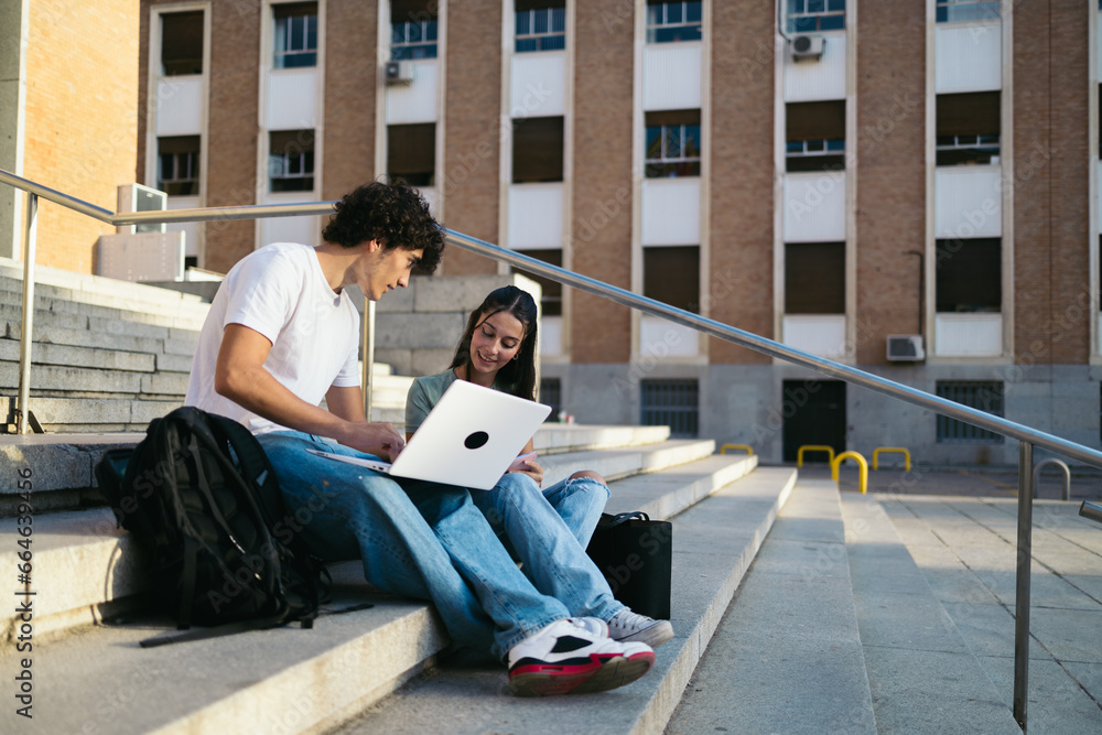 Young college students with casual clothes sitting on stairs outdoors at university and talking while working on laptop and notebook on class project
