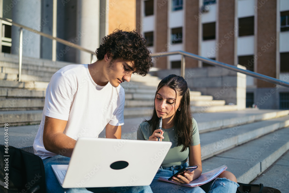 Young college students with casual clothes sitting on stairs outdoors at university and talking while working on laptop on class project Young college students with casual clothes sitting on stairs outdoors at university and talking while working on laptop on class project