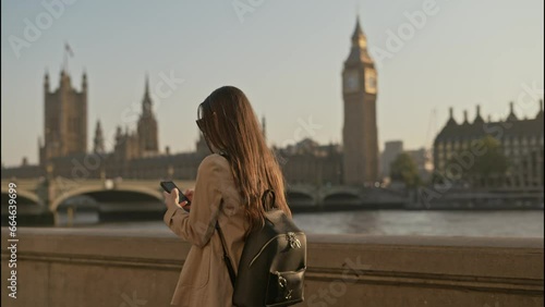Young tourist woman using smartphone while walking along  Thames River with Big Ben Palace of Westminster background view, on a beautiful sunny morning