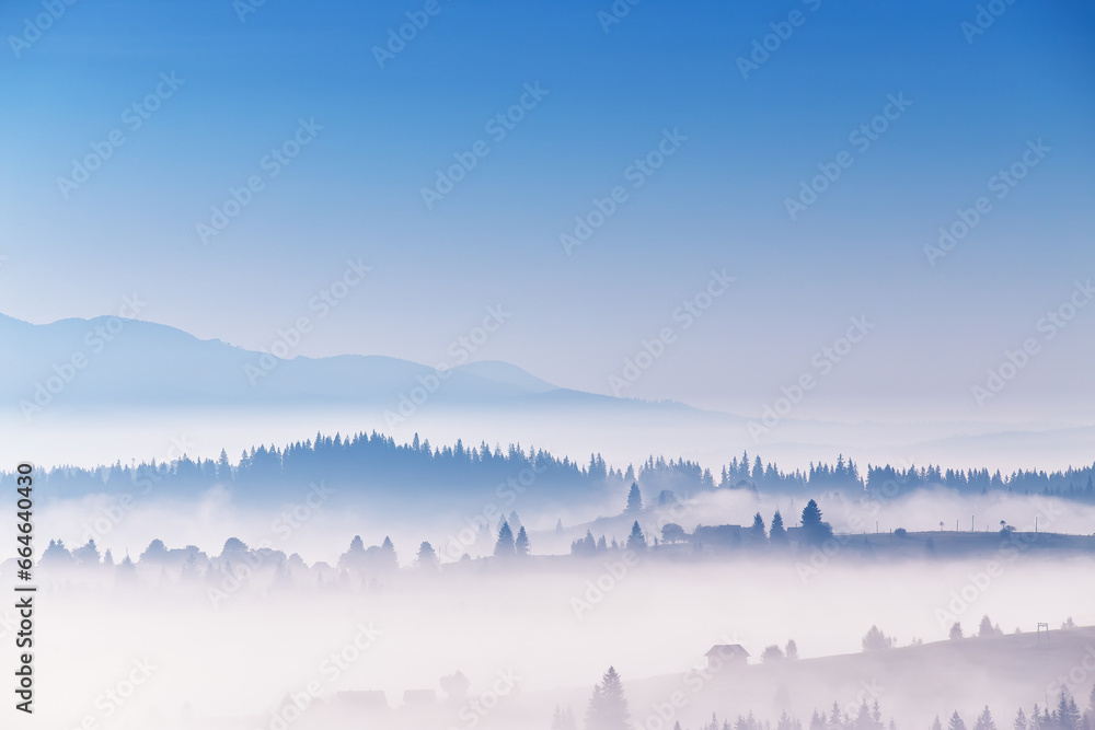 Clear blue sky and clouds of fog moving over alpine village on hills. Tranquil sunrise landscape. Sea of mist in valley. Autumn sunny morning in mountains. Carpathian range