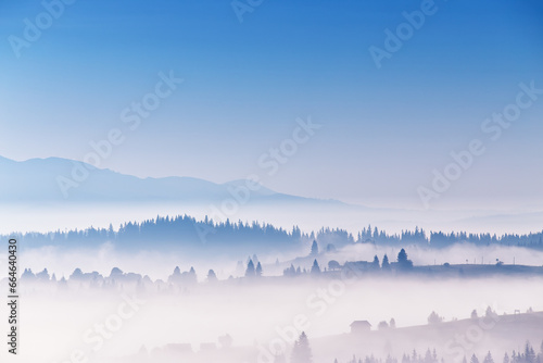 Clear blue sky and clouds of fog moving over alpine village on hills. Tranquil sunrise landscape. Sea of mist in valley. Autumn sunny morning in mountains. Carpathian range
