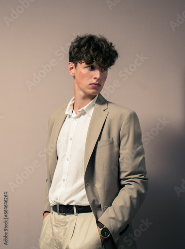 Portrait of a curly-haired teenager in a beige suit and white dress shirt. Beige background. Looking into the floor.