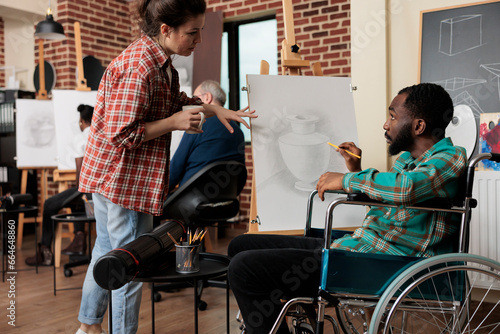 African American disabled guy talking with teacher during group drawing class, improving social skills through collaborative artwork. Teaching art to people with disabilities photo