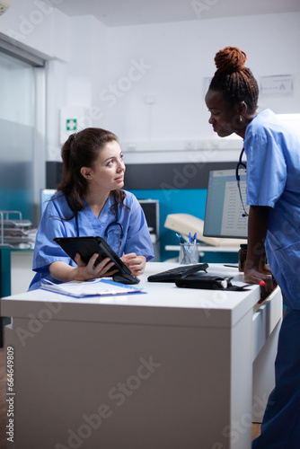 Nurse colleagues in happy relaxed hospital workspace working on tablet in modern professional medical office. Diverse healthcare specialists coworkers in modern clinic using technology