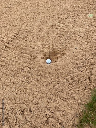 Golf ball plugged in a bunker in the sand next to a golf green also known as greenside. Raked bunker.
