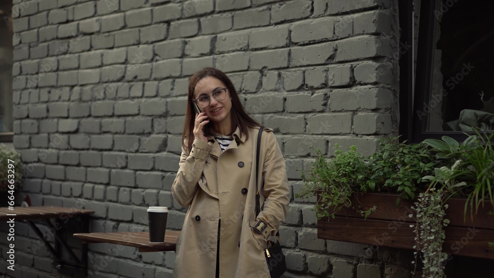 A young woman talks on the phone while standing against a wall on a narrow street. A girl with glasses and a coat speaks on the phone. A young woman talks on the phone while standing against a wall on a narrow street. A girl with glasses and a coat speaks on the phone.