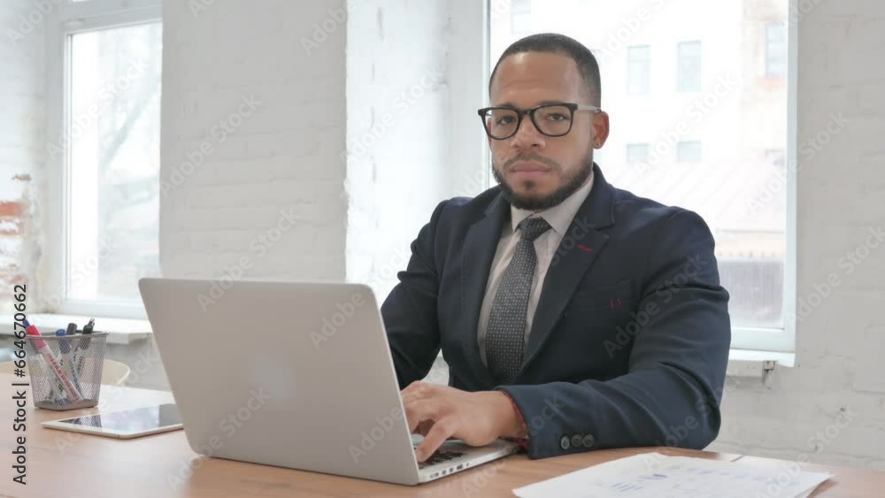 Mixed Race Businessman Looking at Camera while Working on Laptop in Office Mixed Race Businessman Looking at Camera while Working on Laptop in Office
